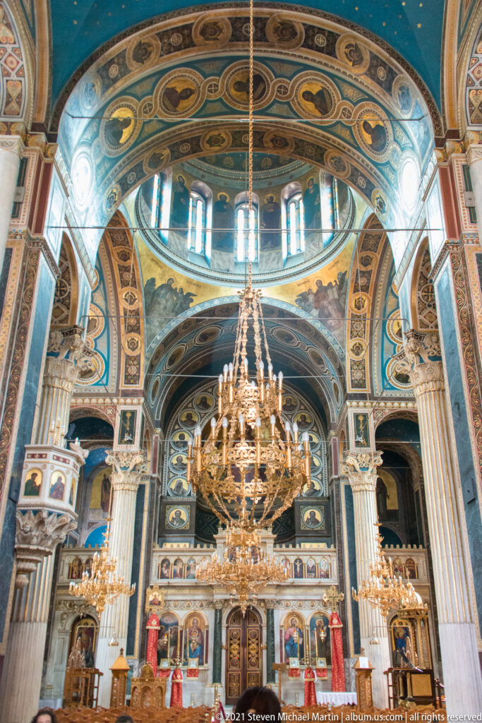Ornate nave of Panagia Chrysospiliotissa with painted vaults, marble columns, large chandeliers, icons, and sanctuary beyond.