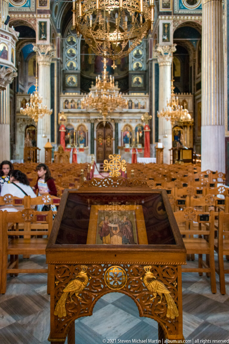 Carved wooden icon stand in the foreground of Panagia Chrysospiliotissa, with chairs, chandeliers, columns, and icons behind.