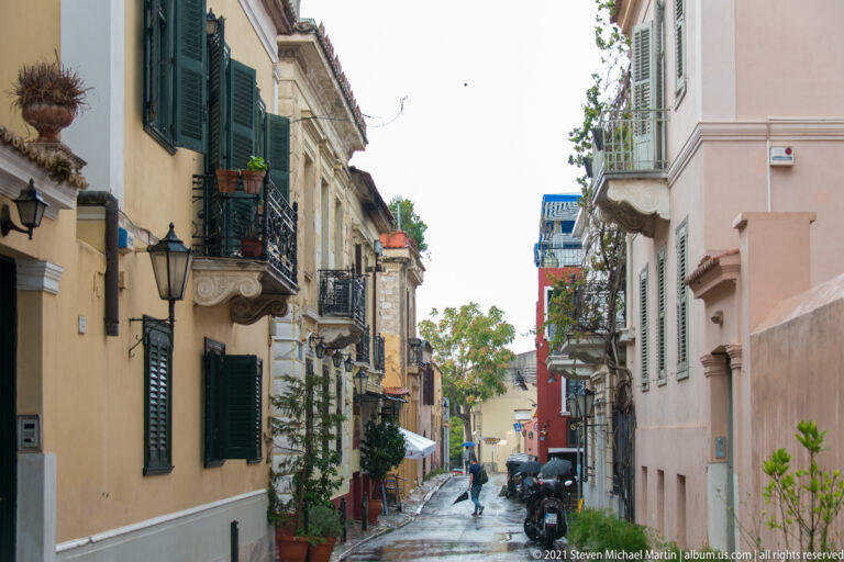 Narrow wet street in Athens lined with pastel houses, iron balconies, shutters, wall lamps, and potted plants.