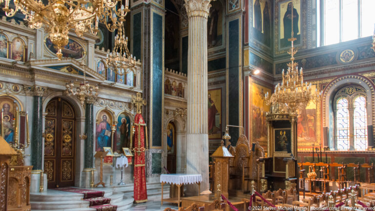 Side aisle of Panagia Chrysospiliotissa with marble column, chandelier, icons, carved doors, altar table, and stained-glass window.