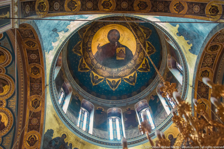 Christ Pantocrator painted in a blue church dome above tall drum windows and surrounding sacred figures.