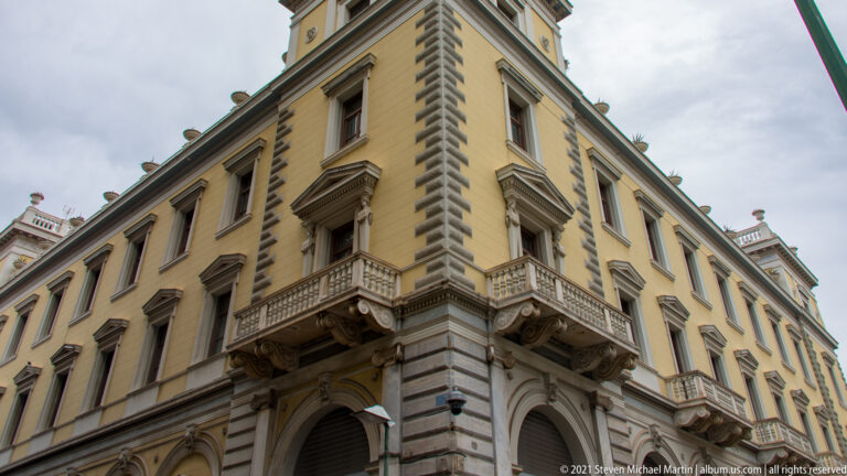 Low-angle corner view of a yellow neoclassical Athens building with arched openings, balconies, and rusticated masonry.