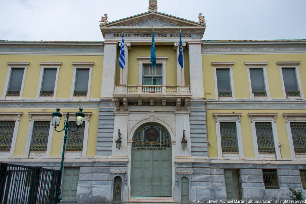 National Bank of Greece façade with central pediment, Greek and European flags, tall windows, and a large green doorway.