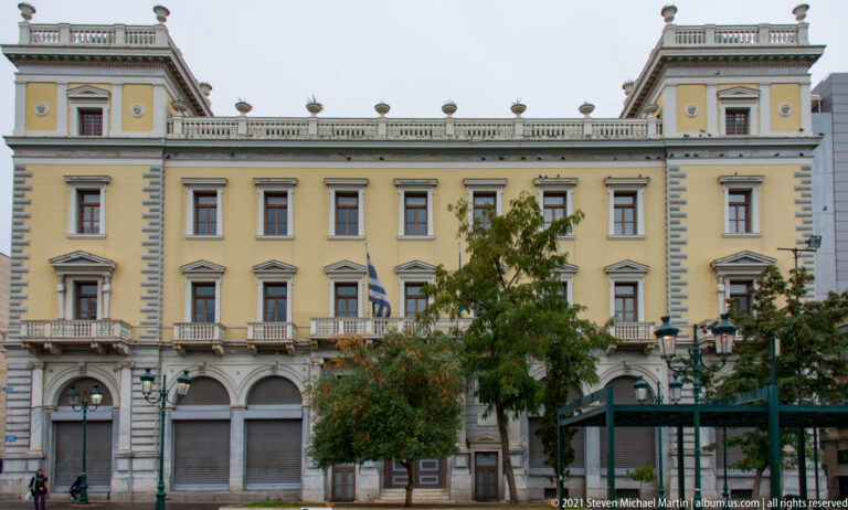 Long yellow neoclassical Athens building with arcaded ground floor, balconies, Greek flags, and trees in front.