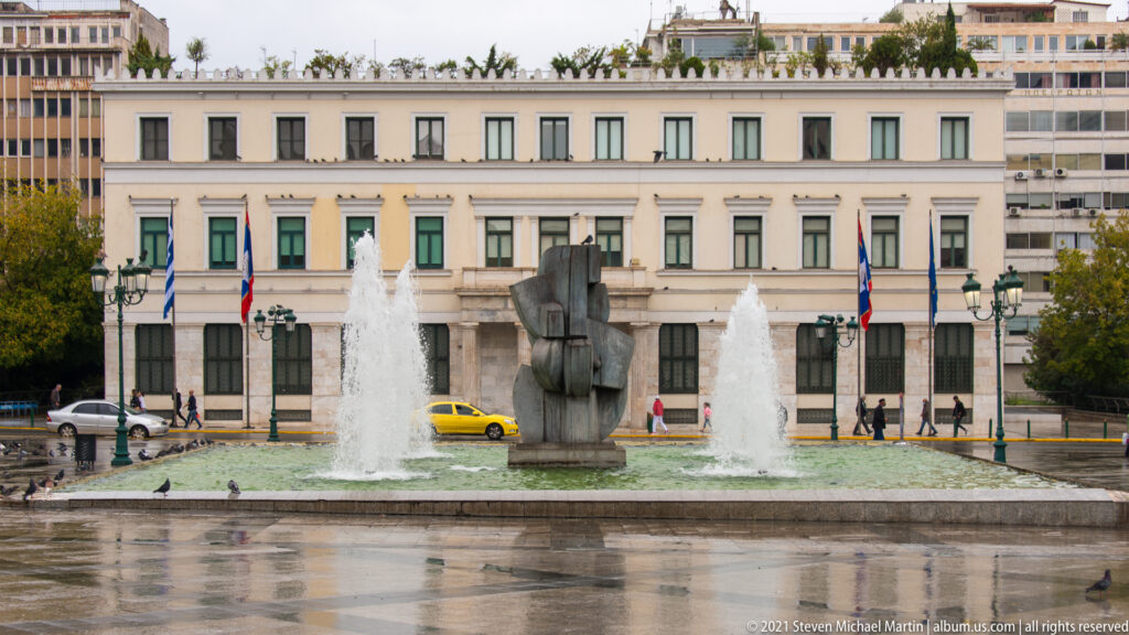 Dark modern sculpture between two fountain jets in an Athens square, with a pale neoclassical building behind.