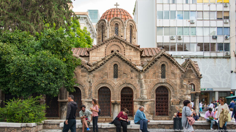 Byzantine Church of Panagia Kapnikarea with tiled roofs, arched windows, dome, visitors, and modern buildings behind.