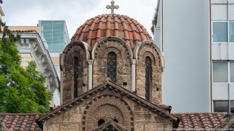 Close view of Panagia Kapnikarea’s red-tiled dome, narrow arched windows, brickwork, stone colonnettes, and cross.