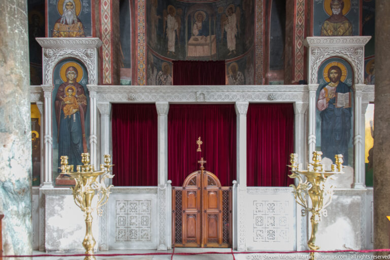 Marble sanctuary screen with wooden doors, red curtains, icons, candle stands, and painted saints inside Panagia Kapnikarea.