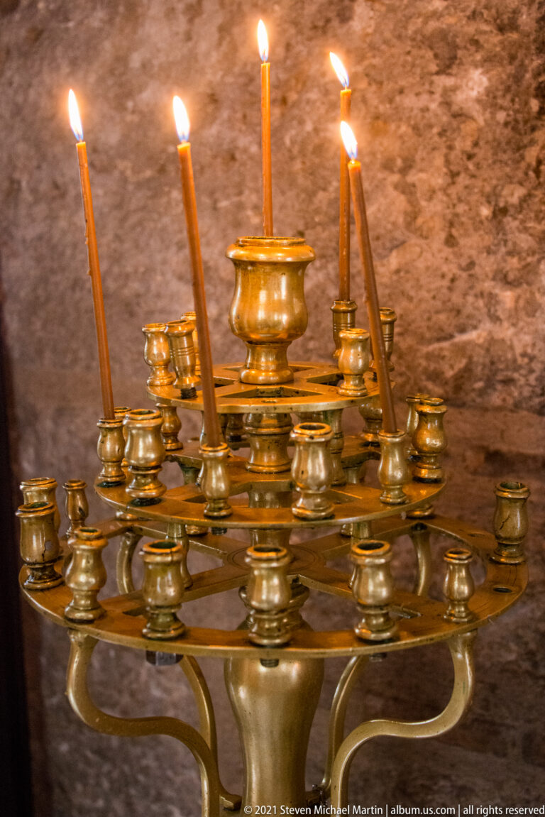 Brass multi-tier church candle stand holding several burning beeswax tapers against a rough stone wall.