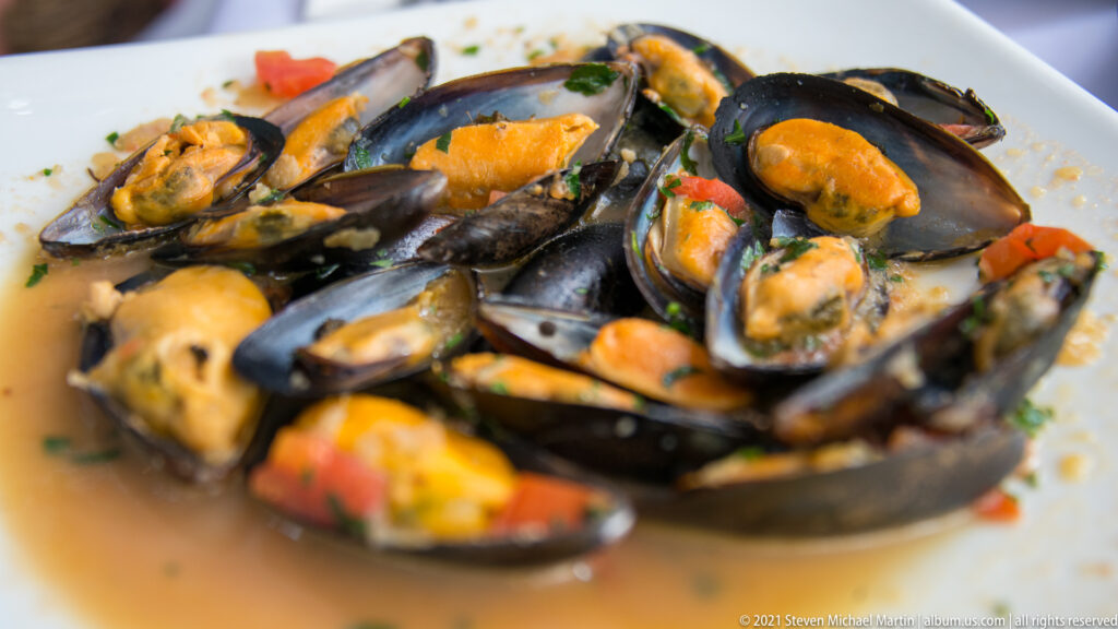 Cooked mussels in dark shells with orange flesh, herbs, diced tomato, and light broth on a white plate.