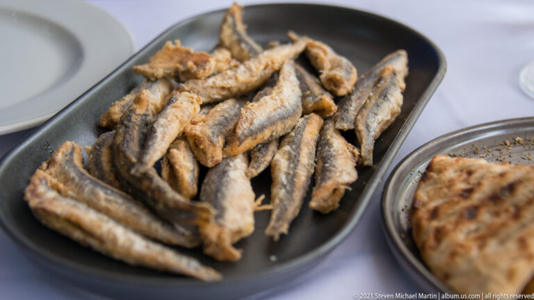 Pile of small whole fried fish with heads and tails on a dark oval restaurant plate.