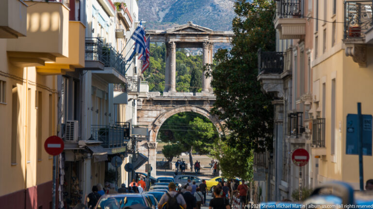 Hadrian's Arch seen at the end of a narrow Athens street lined with buildings, traffic, trees, and pedestrians.