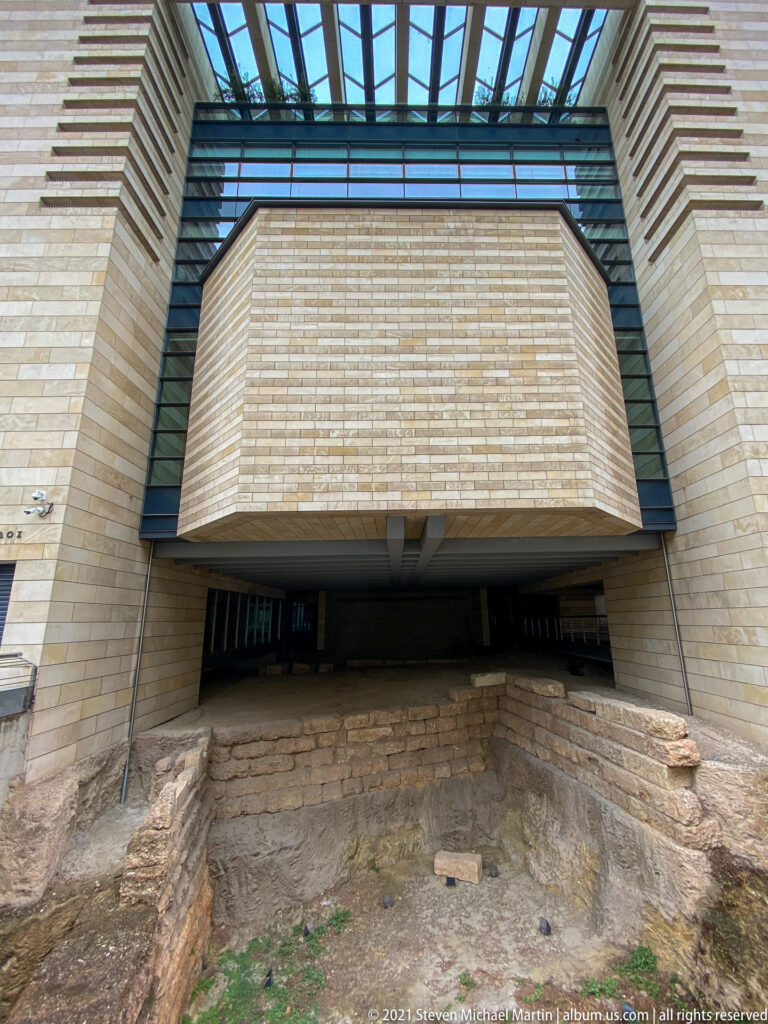 Ancient stone foundations preserved below a modern elevated building with pale block walls and glass structural elements.