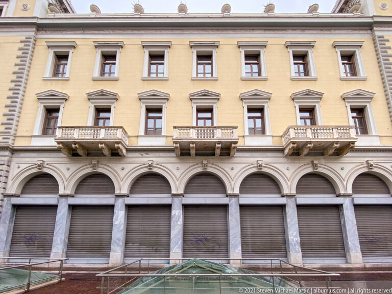 Yellow neoclassical Athens façade with repeated arches, balconies, tall windows, stone pilasters, and classical moldings.