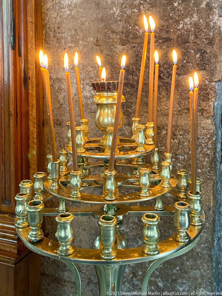 Burning beeswax tapers in a polished brass circular votive candle stand against a rough stone wall.