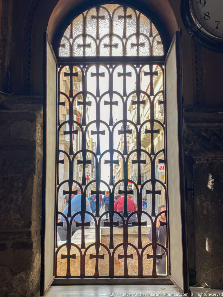 Dark metal cross-pattern grille in an arched church opening, with daylight, pedestrians, and street activity visible beyond.