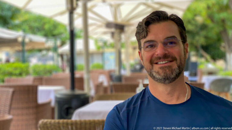 Smiling bearded man in a navy shirt seated at an outdoor restaurant with wicker chairs and greenery behind.
