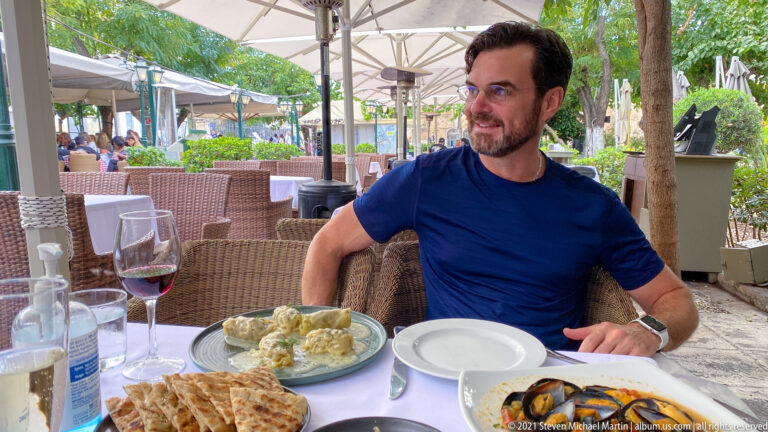 Man at an outdoor Athens restaurant table with bread, wine, water, plates of food, umbrellas, and wicker seating.