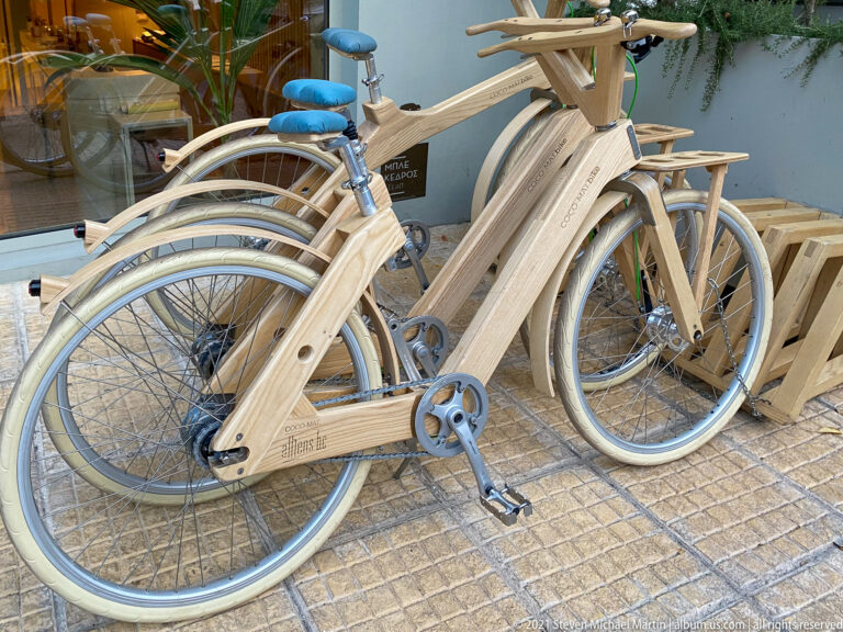 Row of pale wooden-frame bicycles with white tires, metal wheels, rear racks, and blue saddles outside a shop.