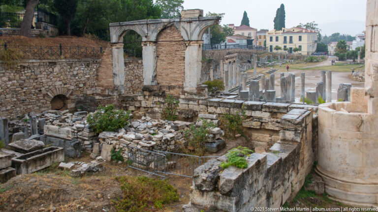 Ruins of the Roman Agora with stone walls, column fragments, standing columns, and later buildings beyond.