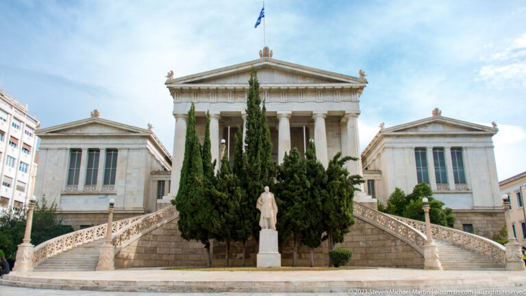 Historic National Library of Greece with Ionic portico, triangular pediment, broad twin staircases, cypress trees, and a foreground statue.