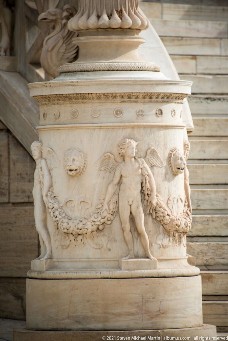 Carved marble pedestal decorated with putti, garlands, rosettes, and lion masks beside the National Library in Athens.