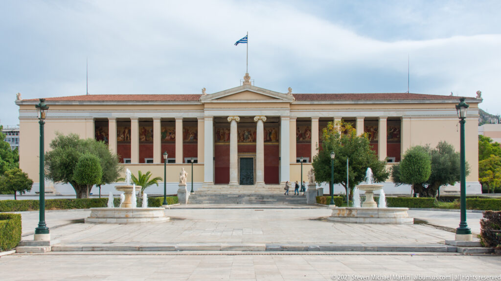 University of Athens neoclassical façade with central Ionic portico, red wall panels, Greek flag, fountains, and landscaped forecourt.