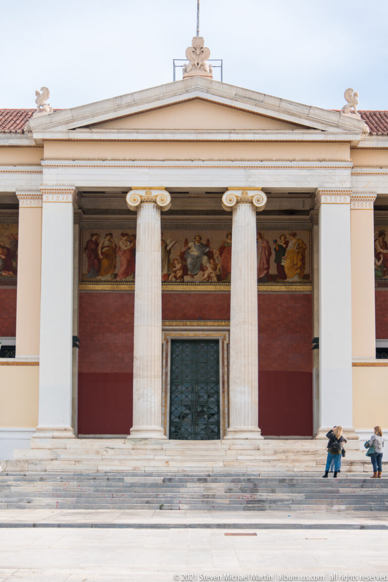 Central University of Athens portico with tall Ionic columns, broad steps, dark doors, red walls, and a painted frieze.