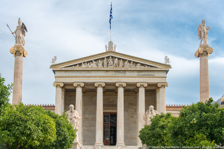 Academy of Athens façade with Ionic portico, sculpted pediment, two tall statue-topped columns, trees, and marble figures.