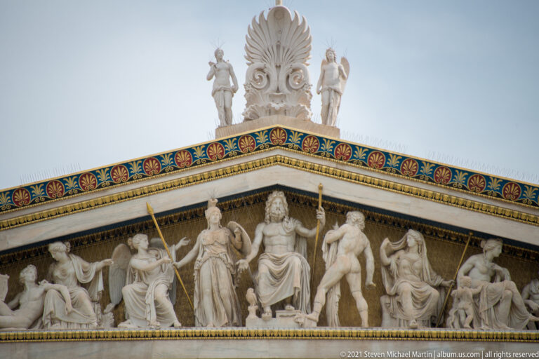 Marble pediment sculptures at the Academy of Athens beneath a brightly colored palmette cornice and roof ornaments.