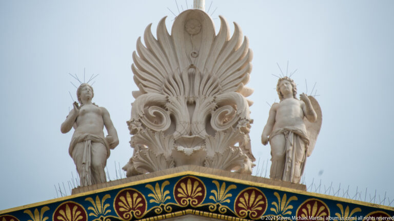 Large marble palmette roof ornament flanked by two statues above the brightly colored cornice of the Academy of Athens.