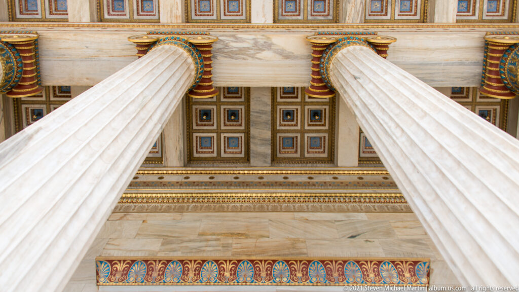 Upward view between fluted Academy of Athens columns toward a richly painted geometric coffered ceiling.
