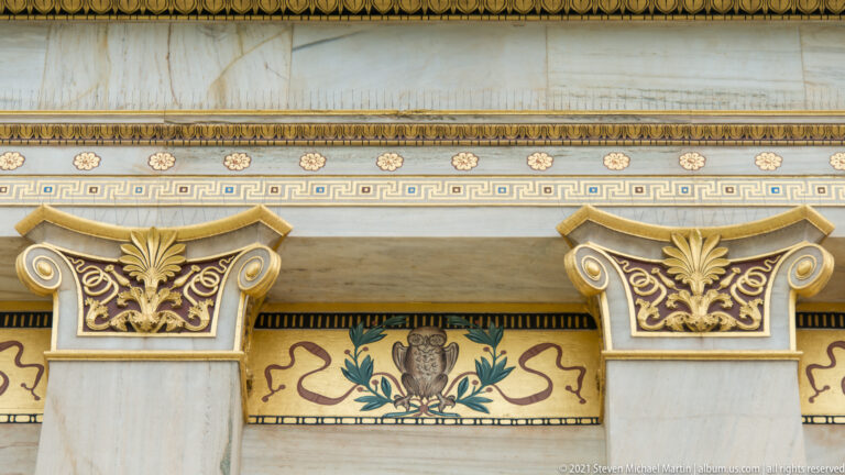 Painted owl and foliage motif between marble pilasters beneath Greek-key and gilded moldings at the Academy of Athens.