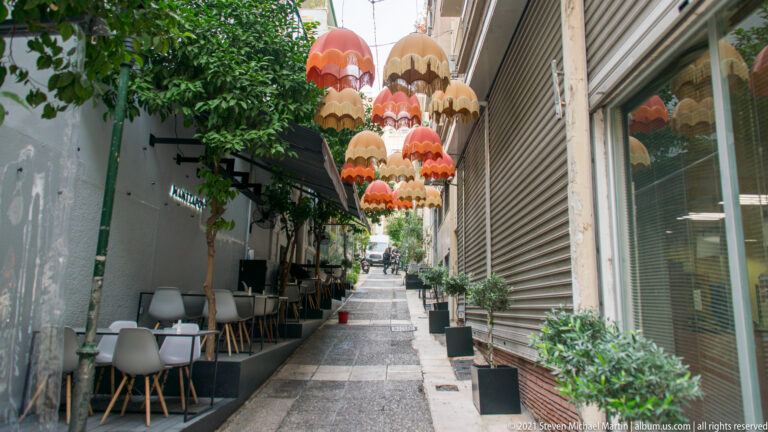 Narrow Athens alley lined with café tables and plants beneath clusters of red and cream hanging scalloped lampshades.