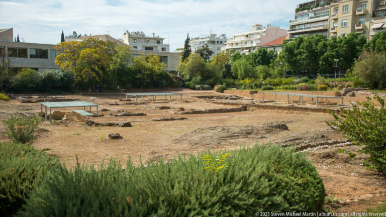Low excavated stone foundations at the Lykeion site with protective shelters, trees, shrubs, and apartment buildings beyond.