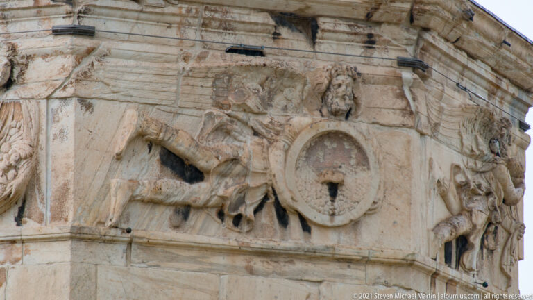 Winged relief figure beside a circular sundial and gnomon on the marble exterior of the Tower of the Winds.