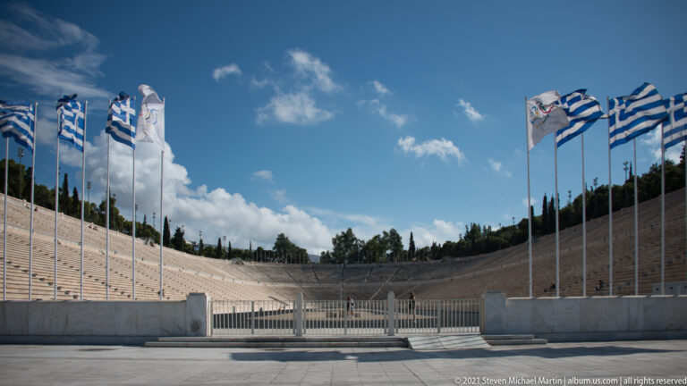 Panathenaic Stadium with long central track, pale marble seating, and rows of Greek flags beneath a blue sky.