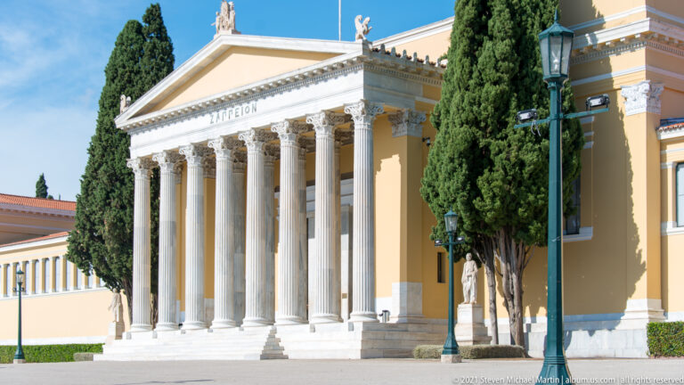Oblique view of the Zappeion with white Corinthian columns, yellow walls, triangular pediment, and tall cypress trees.