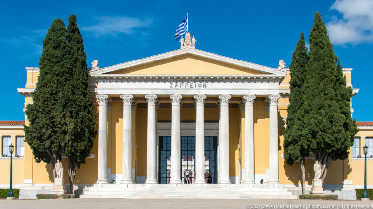 Frontal Zappeion façade with Corinthian portico, triangular pediment, cypress trees, statues, and Greek flag.