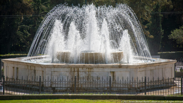 Circular stone fountain with high central sprays and arcing water jets in the shaded Zappeion Gardens.
