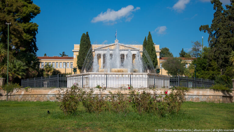Garden fountain with rose beds and lawn in front of the yellow Zappeion building and tall cypress trees.