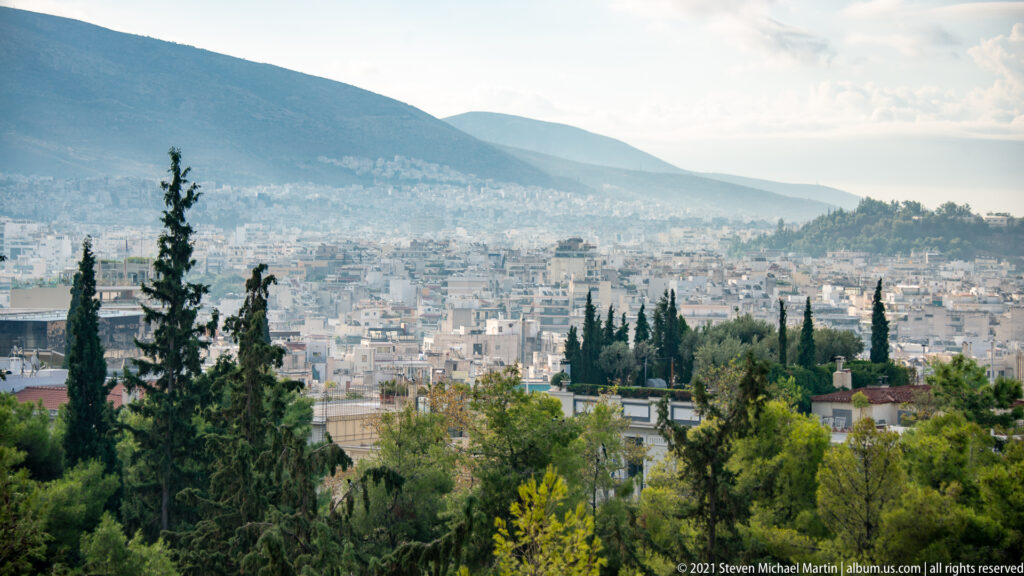 Dense Athens cityscape seen from the Acropolis, framed by cypress trees with a mountain ridge in the distance.
