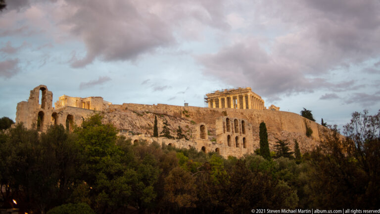 Wide evening view of the Acropolis with the Parthenon lit above long stone walls, trees, and south-slope ruins.