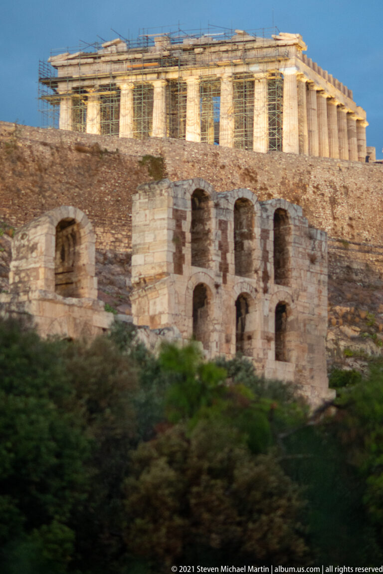 Parthenon lit above the Acropolis wall, with the arched Roman stage building of the Odeon in front.