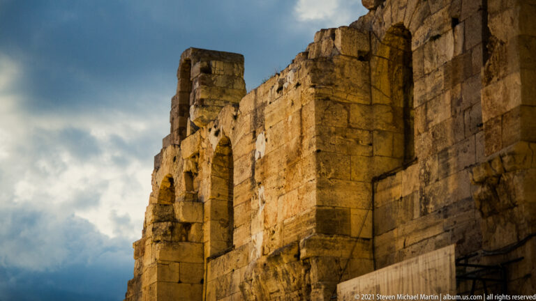 Low-angle close view of massive weathered stone and brick walls of the Odeon against dark clouds.