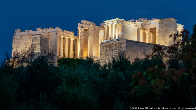 Night view of the illuminated Propylaia and Temple of Athena Nike above dark trees on the Acropolis.