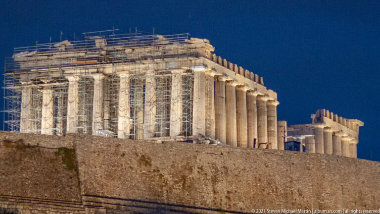 Night view of the Parthenon with bright floodlighting and extensive scaffolding around its Doric columns.