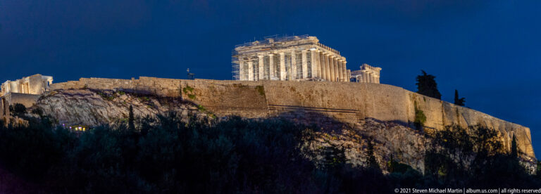 Illuminated Parthenon above long Acropolis walls and a dark rocky slope under a deep blue night sky.