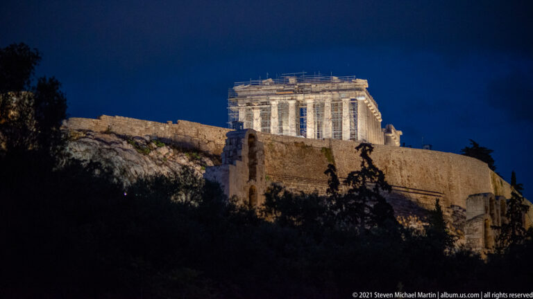 Distant night view of the brightly illuminated Parthenon above the dark Acropolis rock and walls.