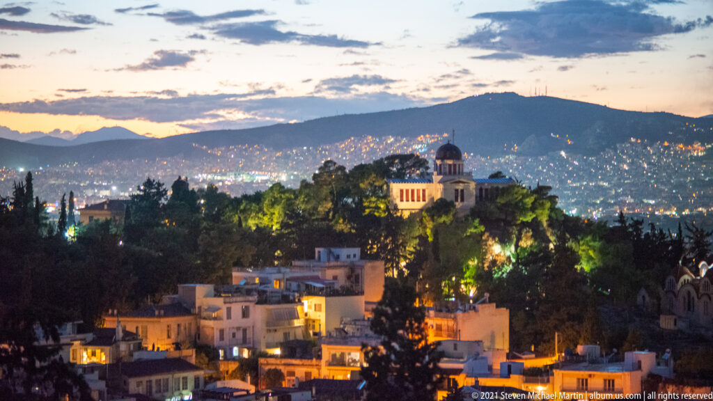 Dusk cityscape of Athens with a domed church among trees, dense lights, and a dark mountain ridge beyond.